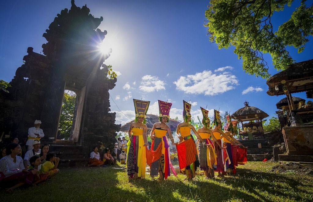 Danse traditionnelle balinaise dans un temple hindou, avec des danseuses en costumes colorés et le soleil brillant à travers les arbres.