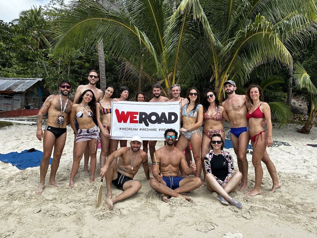 Un groupe de voyageurs WeRoad souriants posant sur une plage tropicale aux Philippines avec le drapeau de l'entreprise sous des palmiers.