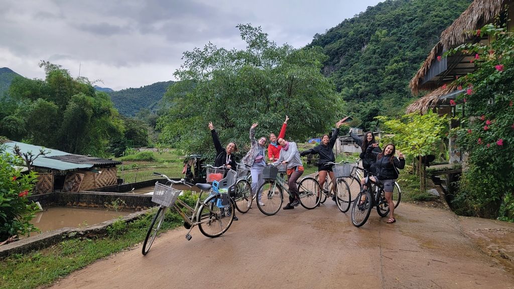 Groupe de voyageurs posant avec leurs vélos dans un village traditionnel entouré de nature luxuriante.