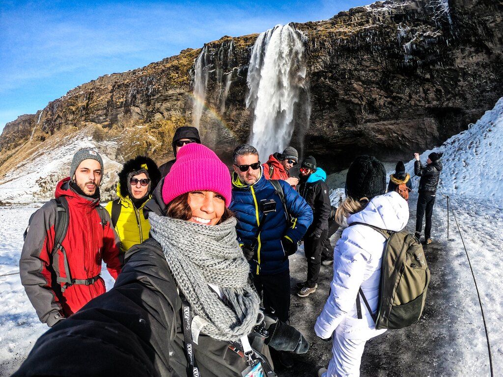 Groupe de voyageurs WeRoad posant devant la cascade Seljalandsfoss, entourée de neige et avec un arc-en-ciel en arrière-plan, en Islande.