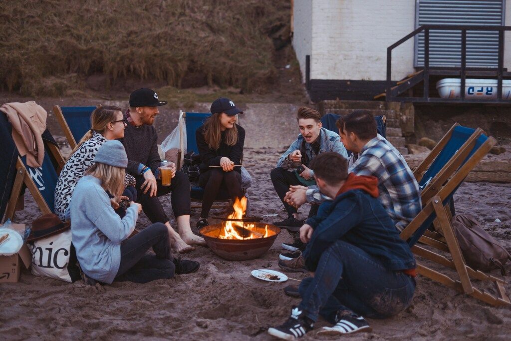 Jeunes réunis autour d’un feu de camp sur une plage, discutant et profitant d’un moment convivial en soirée.