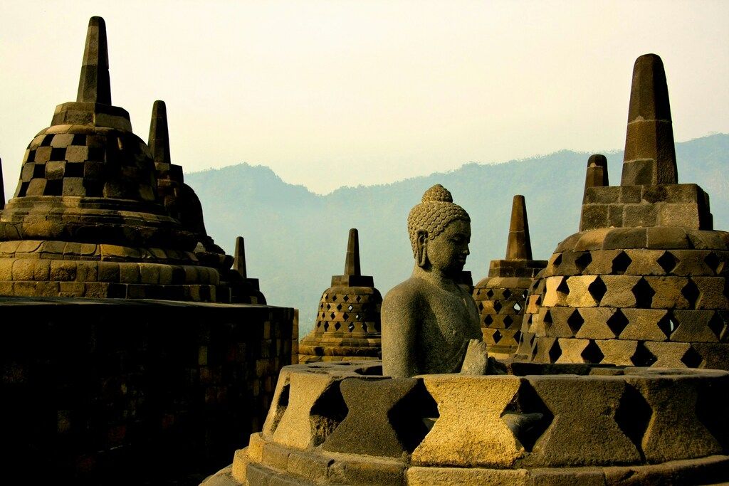 Statue de Bouddha entourée de stupas en pierre au temple de Borobudur, avec les montagnes en arrière-plan.