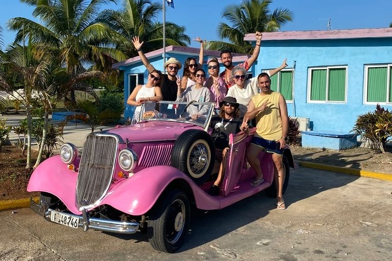 Groupe de voyageurs WeRoad souriants à Cuba dans une voiture décapotable rose vintage, avec des palmiers et une maison bleue en arrière-plan.