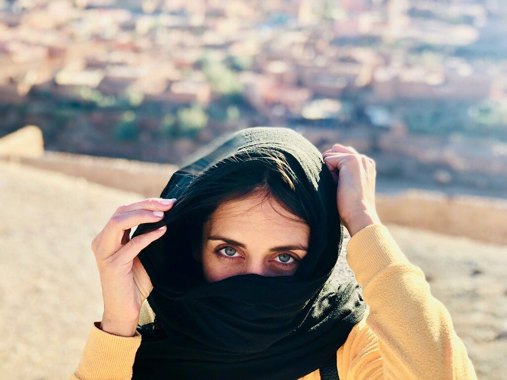 Femme aux yeux bleus levant un foulard noir sur la tête pour couvrir son visage, avec une ville floue en arrière-plan.