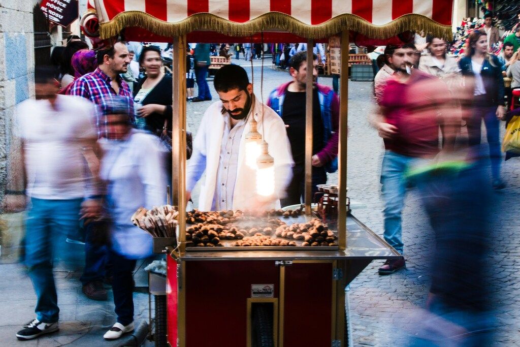 Vendeur de marrons chauds debout derrière un chariot à rayures rouges et blanches, entouré de passants en mouvement dans une rue animée.