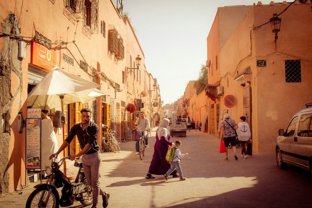 Ruelle étroite d’une médina aux murs ocre, où un jeune homme pousse une mobylette sous un parasol et une femme voilée traverse la rue avec un enfant, tandis que d’autres passants se dispersent.