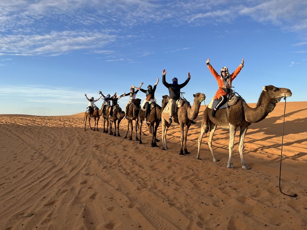 Caravane de touristes montant chacun un chameau dans le désert, alignés sur le sable rouge et levant les bras.