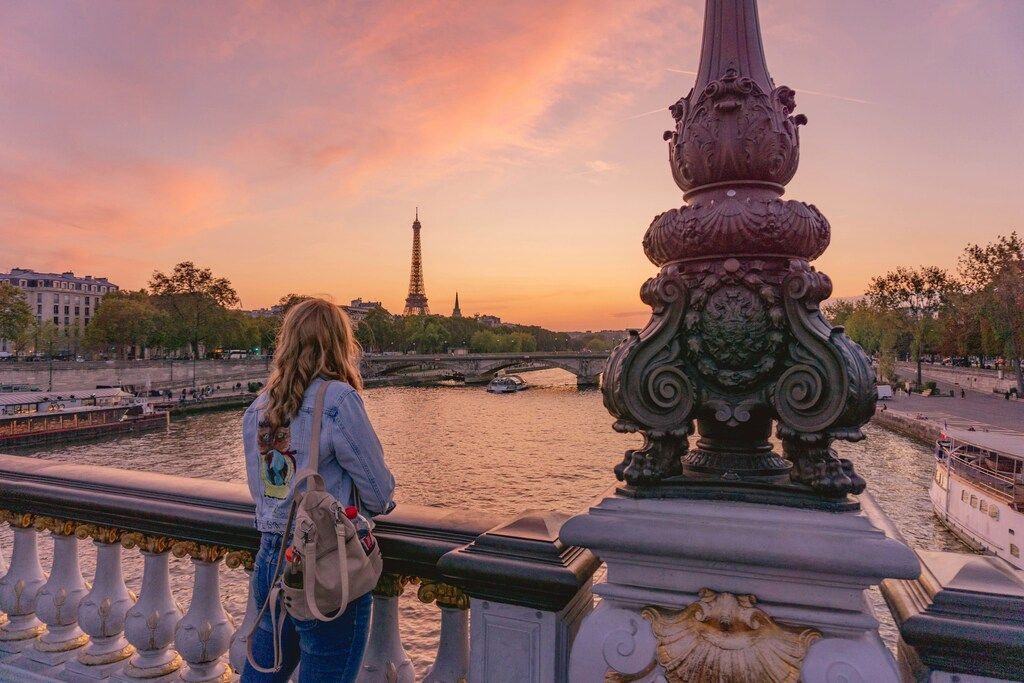 Femme regardant la tour Eiffel depuis le pont Alexandre III au coucher du soleil, avec la Seine en contrebas.