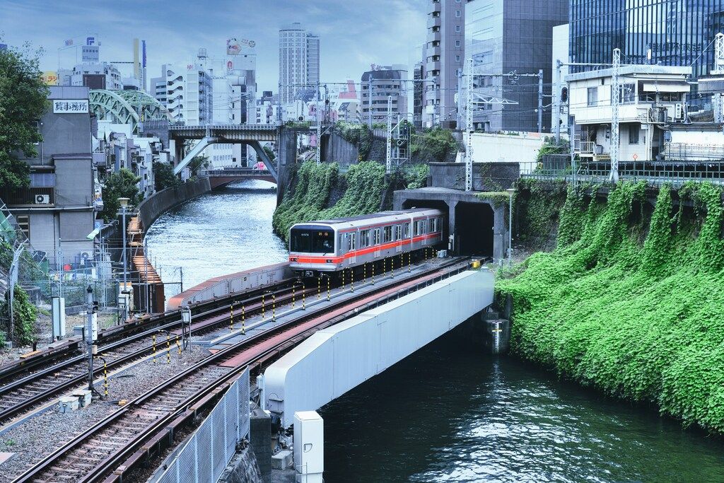 Rame de métro sortant d’un tunnel le long d’une rivière au cœur de Tokyo, entourée d’immeubles et de végétation.