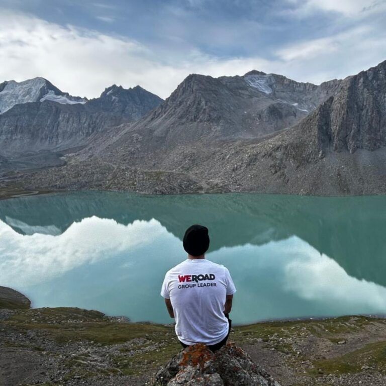 Un coordinateur WeRoad assis face au lac Ala Kul, entouré par les montagnes bleues du Tien Shan au Kirghizistan.