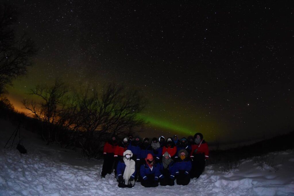 Un groupe de voyageurs Weroad pose dans la neige sous une aurore boréale verte et un ciel étoilé en Suède.
