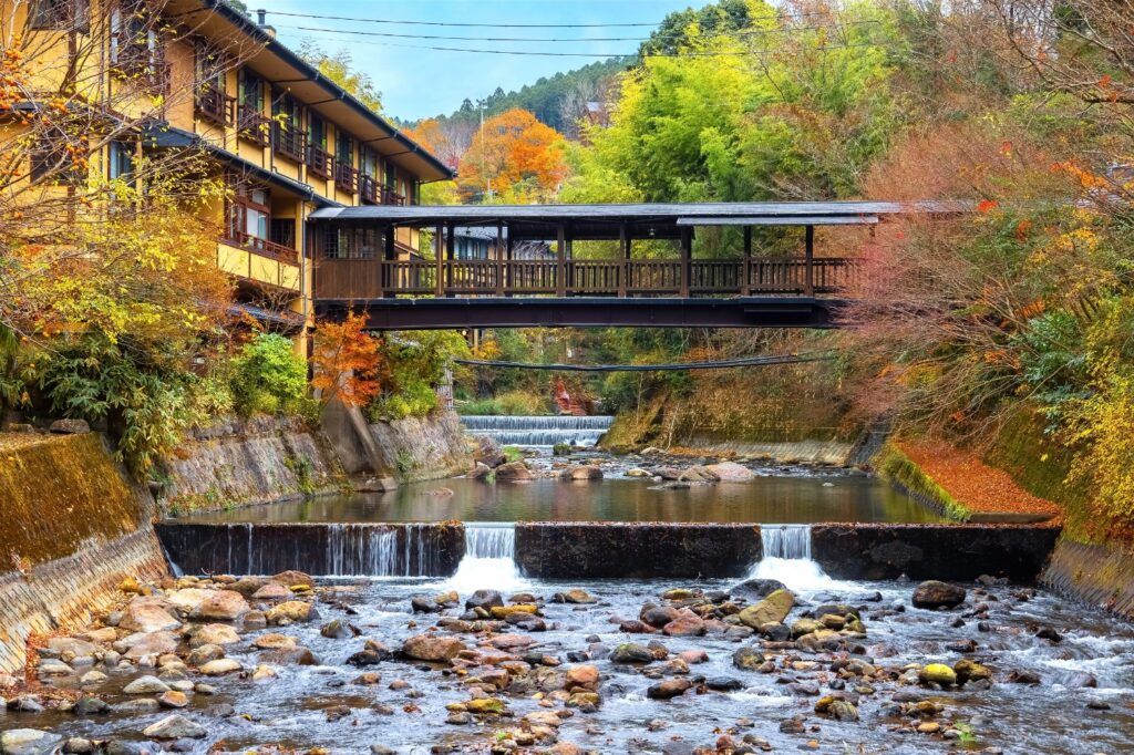 Passerelle reliant un ryokan aux Kurokawa Onsen au Japon, avec une rivière et de la végétation automnale au premier plan.