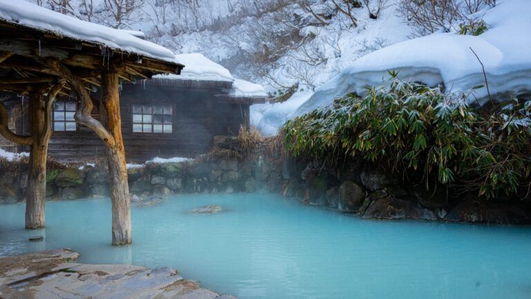 Un onsen (bain chaud) extérieur aux eaux turquoise dans un paysage enneigé, avec des cabanes traditionnelles en bois