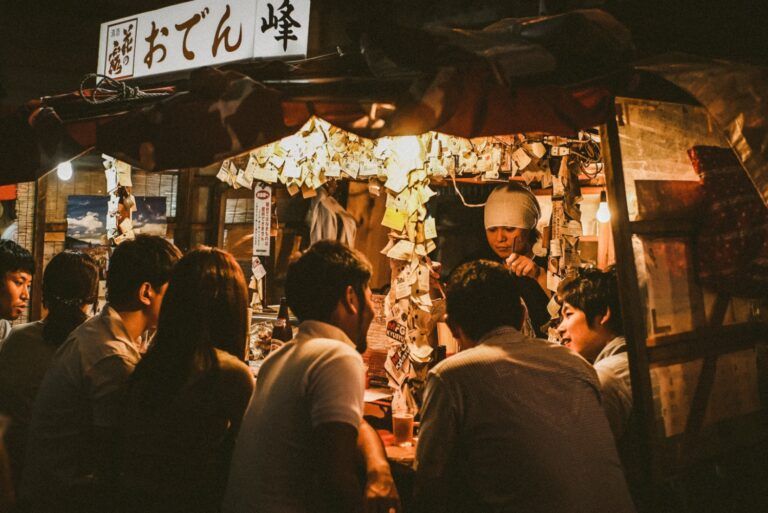 Stand de yatai traditionnel japonais, éclairé la nuit, où des clients sont assis au comptoir et discutent avec le chef sous une enseigne d'Oden.
