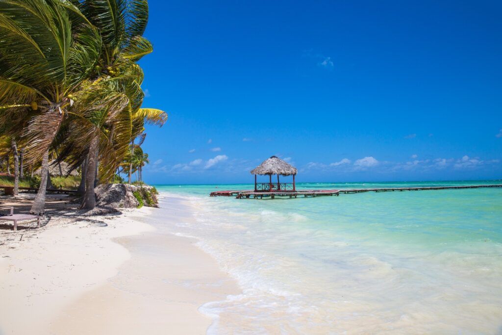 Vue idyllique de Cayo Guillermo : jetée en bois avec gazebo au toit de chaume sur une mer turquoise peu profonde et du sable blanc.