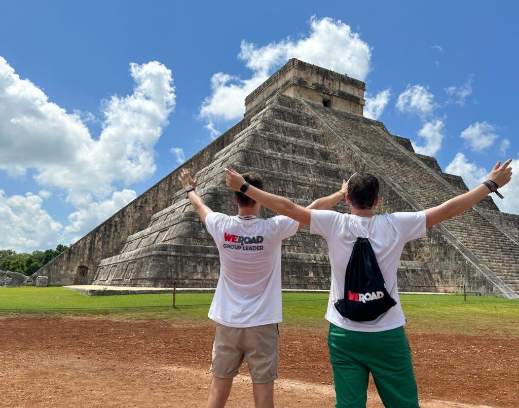 Deux WeRoad Group Leaders vus de dos, les bras levés devant la pyramide monumentale de Chichén Itzá sous un ciel bleu parsemé de nuages blancs.