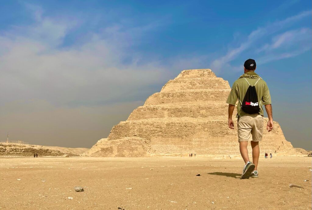 Un voyageur avec un sac à dos WeRoad marche vers une pyramide à degrés dans le désert égyptien.