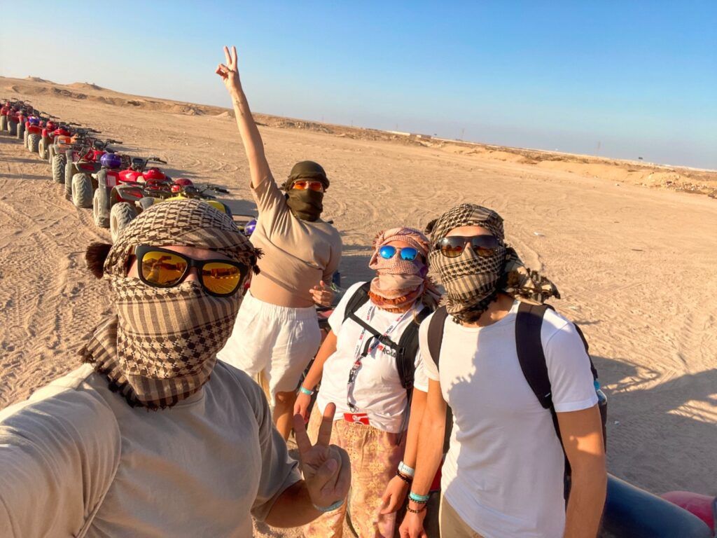 Un groupe de voyageurs WeRoad avec des chèches et des lunettes de soleil pose devant une file de quads dans le désert égyptien.