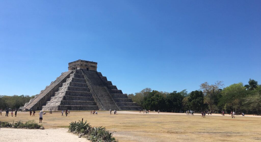 L'imposante pyramide de Kukulcán à Chichén Itzá, au Mexique, s'élevant sous un ciel bleu pur, entourée de nombreux visiteurs sur une vaste esplanade ensoleillée.