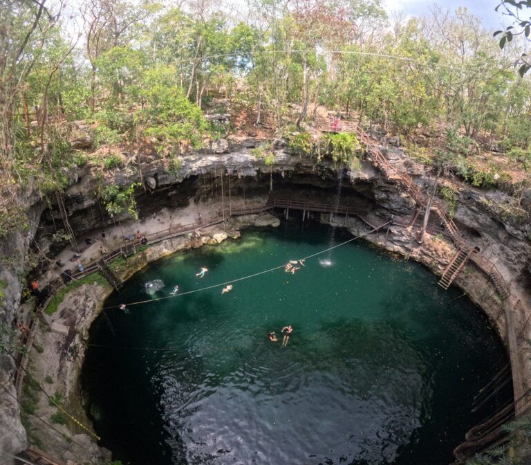 Le cénote de Miguel Colorado au Mexique, montrant des voyageurs nageant et faisant de la tyrolienne au-dessus d'une eau vert émeraude entourée de parois rocheuses et d'une forêt dense.