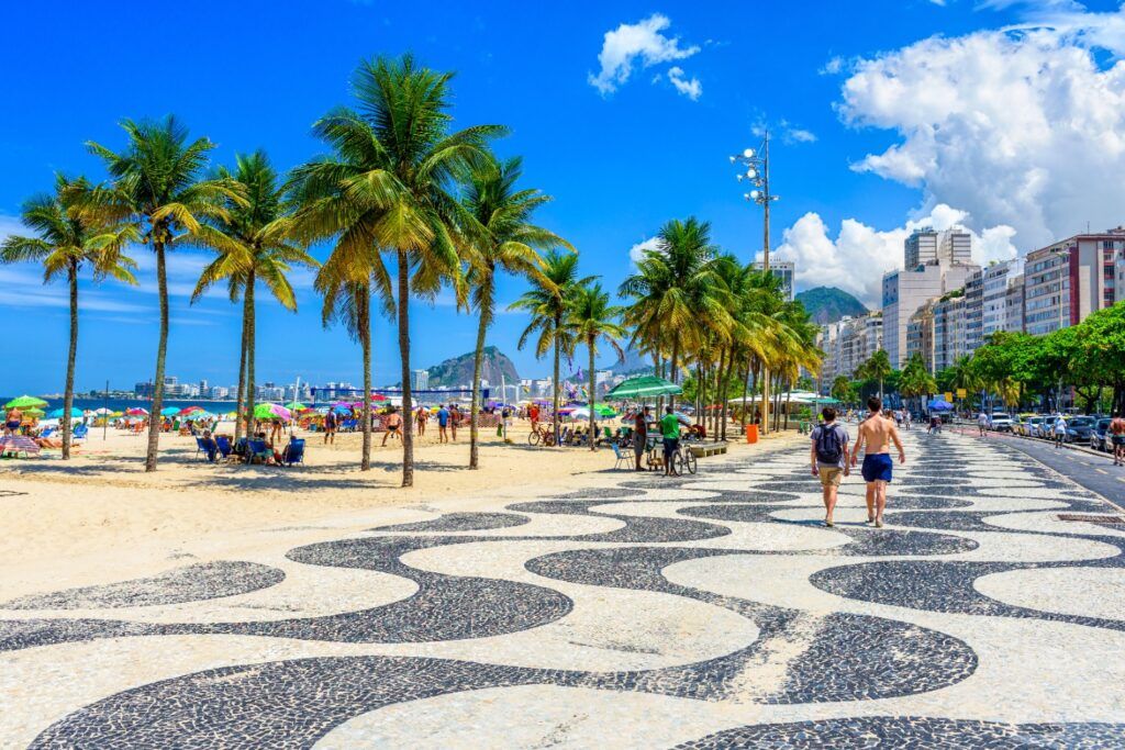 Promenade de Copacabana avec ses mosaïques emblématiques, bordée de palmiers et de la plage sous un ciel bleu à Rio de Janeiro.