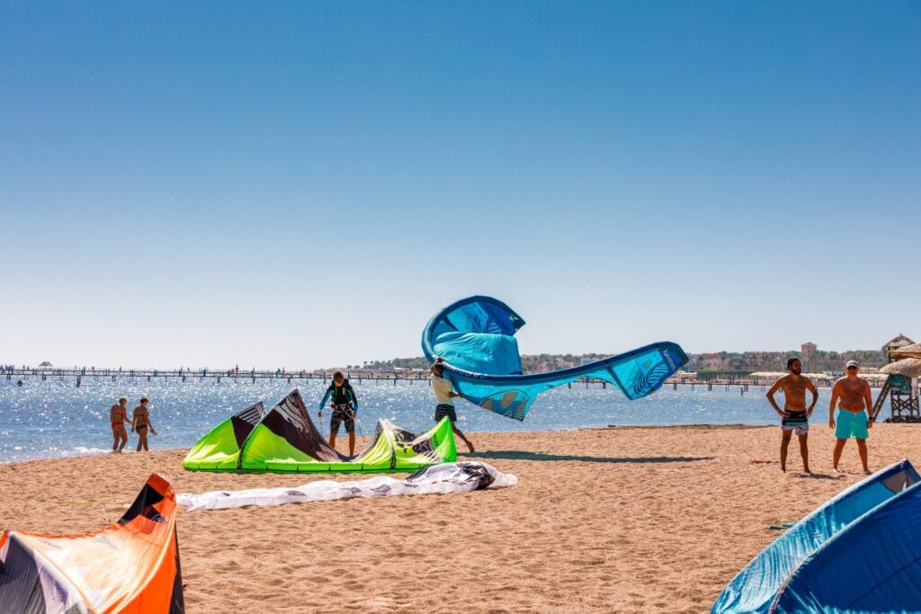 Des personnes préparent des voiles de kitesurf sur une plage de sable en Égypte, au bord d'une mer bleue scintillante.