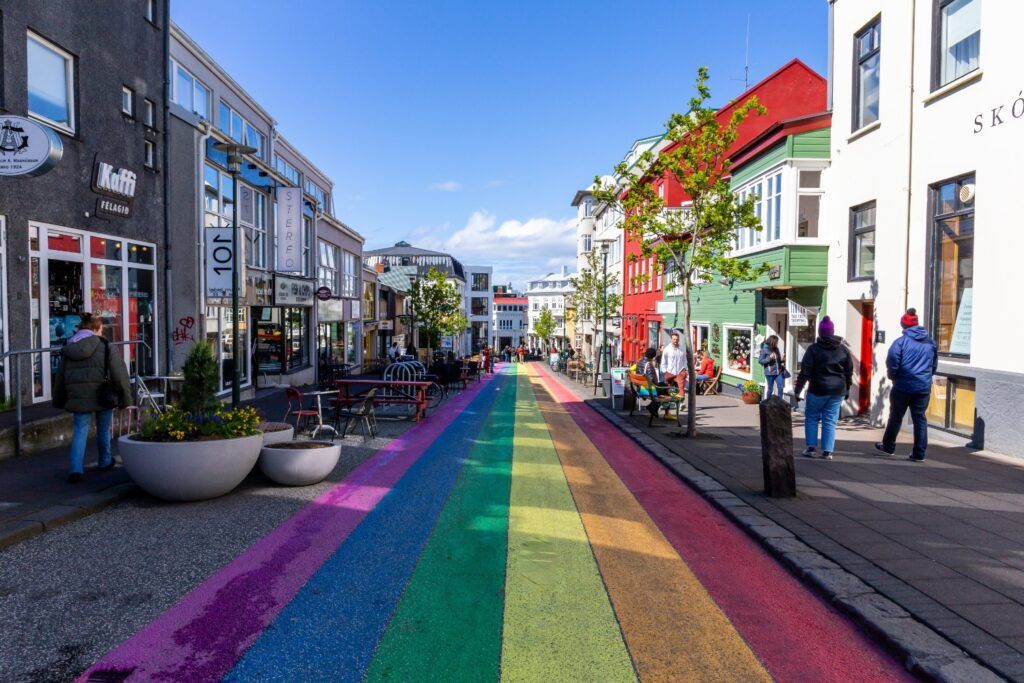 Rue piétonne pavée à Reykjavik avec une grande bande arc-en-ciel peinte au sol, bordée de bâtiments colorés et de boutiques.