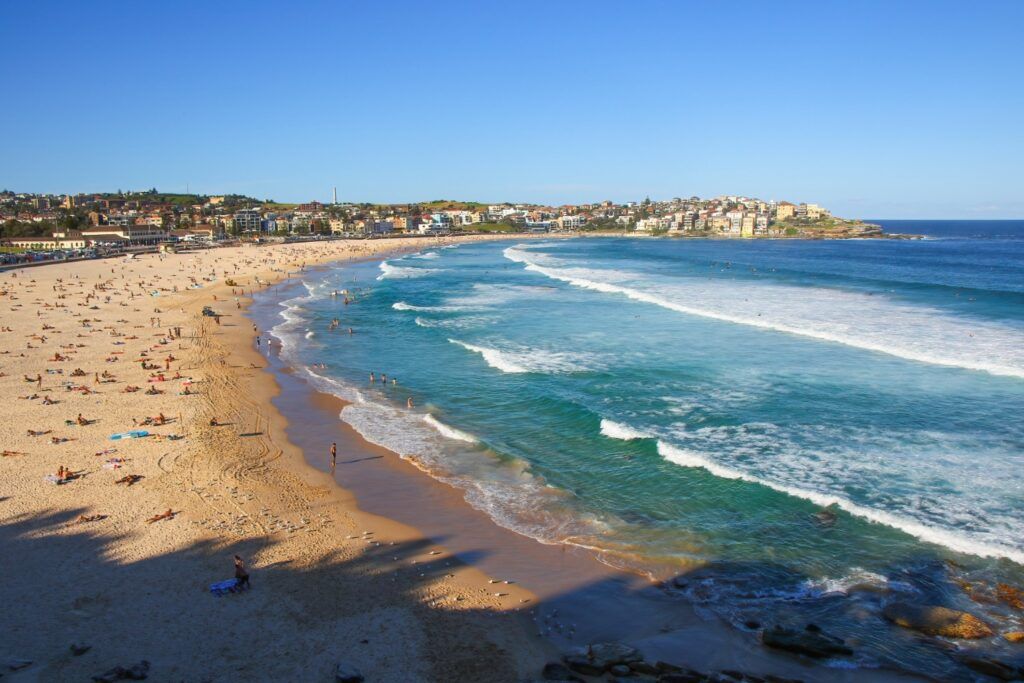 Vue de la plage de Bondi en Australie, avec de nombreux baigneurs sur le sable et des surfeurs dans les vagues sous un ciel bleu.