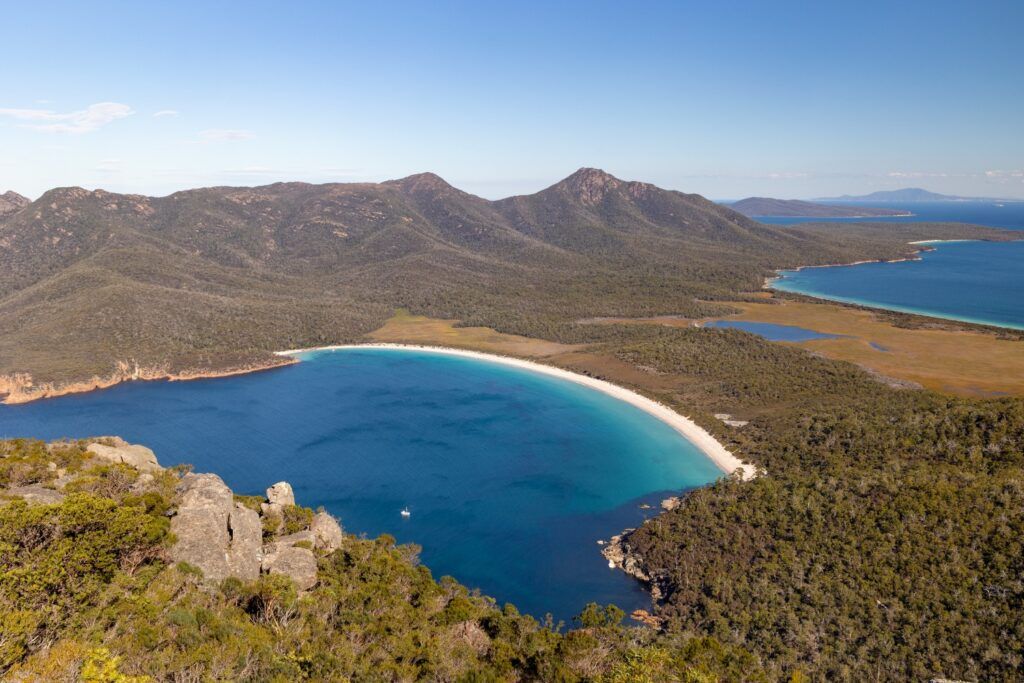 Vue panoramique de la courbe parfaite de sable blanc de Wineglass Bay en Tasmanie, entourée de montagnes et de forêts.