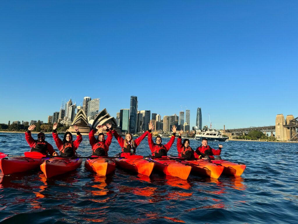 Un groupe de voyageurs WeRoad en kayak rouge sur l'eau à Sydney, avec l'Opéra et le Harbour Bridge en arrière-plan.
