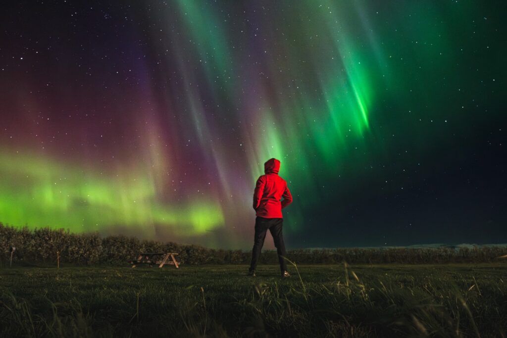 Une personne en veste rouge observe une aurore boréale spectaculaire aux teintes vertes et violettes dans un ciel étoilé.