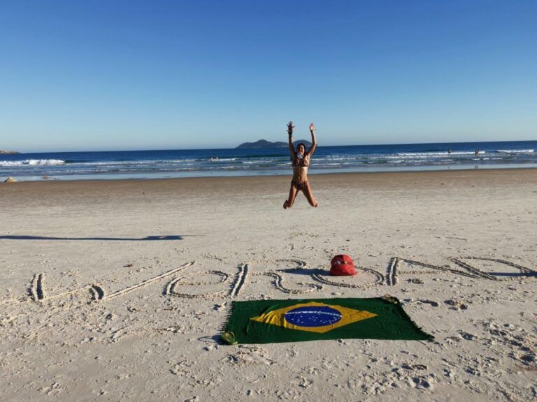 Un voyageur sautant de joie sur une plage de sable blanc au Brésil, avec 'WeRoad' écrit dans le sable et un drapeau brésilien déployé.