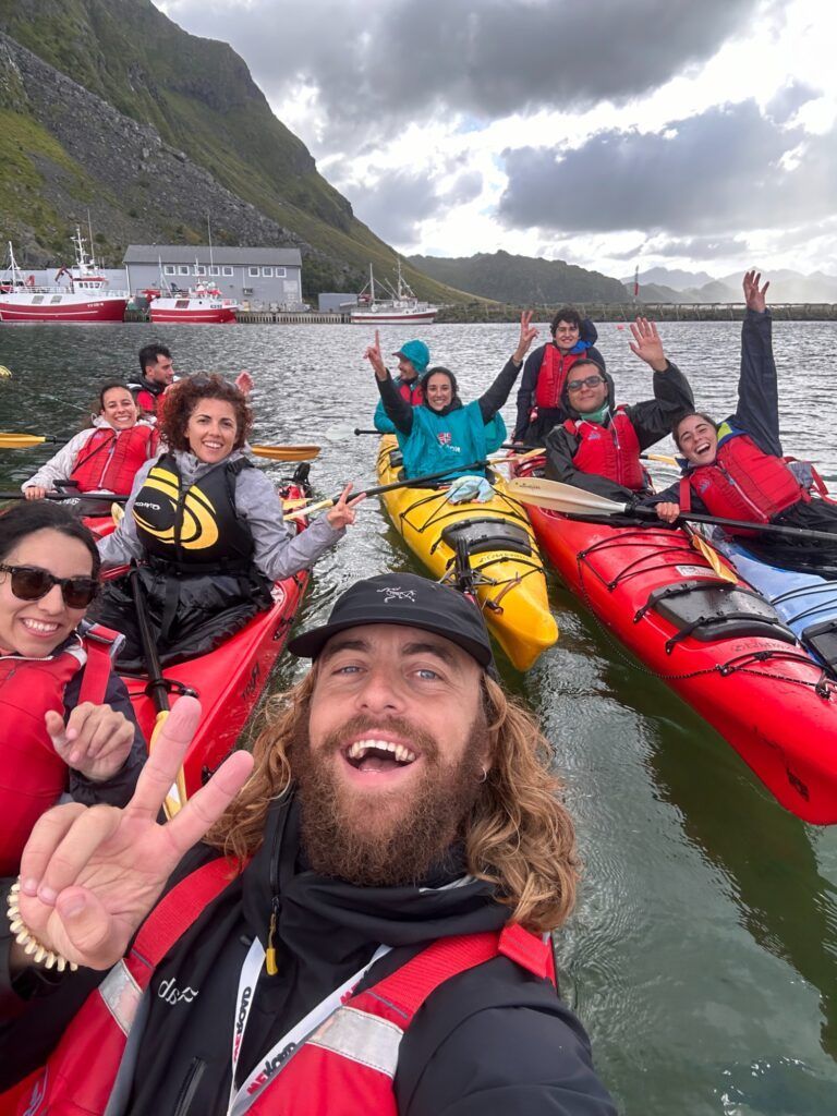 Un groupe de voyageurs WeRoad joyeux en kayak sur un fjord norvégien, souriant pour un selfie sous un ciel nuageux.