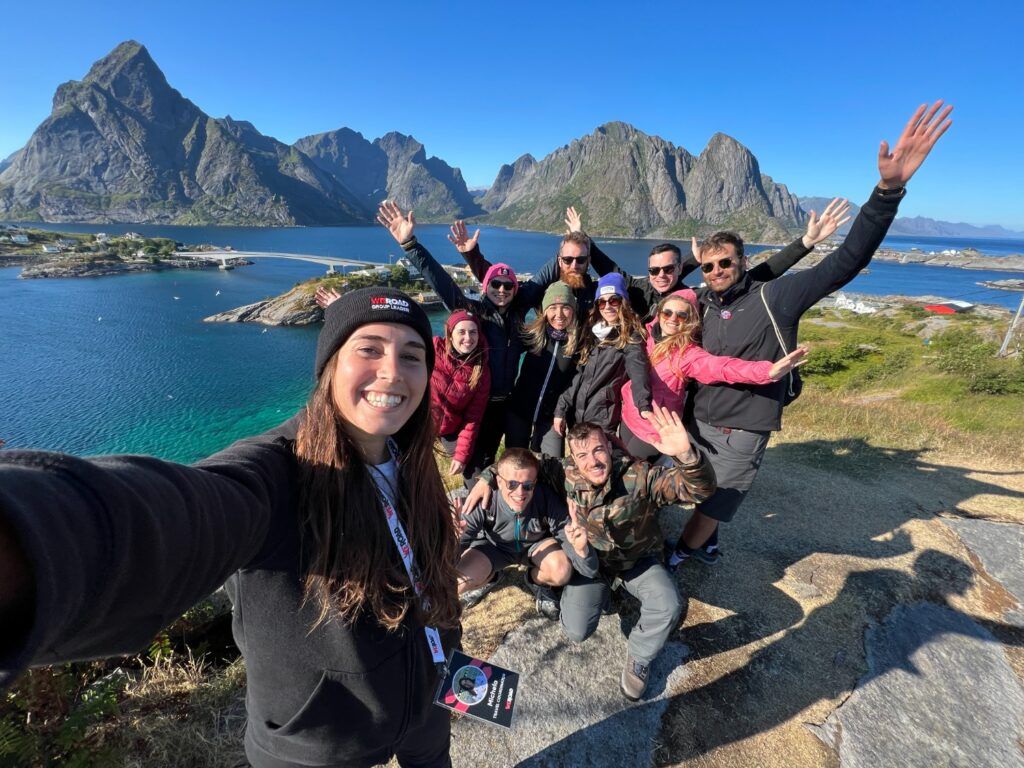 Un groupe de voyageurs WeRoad prend un selfie souriant devant les montagnes majestueuses et les eaux turquoise des îles Lofoten en Norvège.
