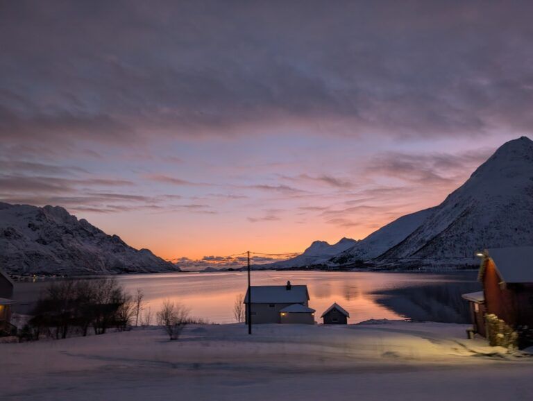Un paysage hivernal paisible en Norvège avec des maisons enneigées au bord d'un fjord sous un ciel de coucher de soleil violet et orange.