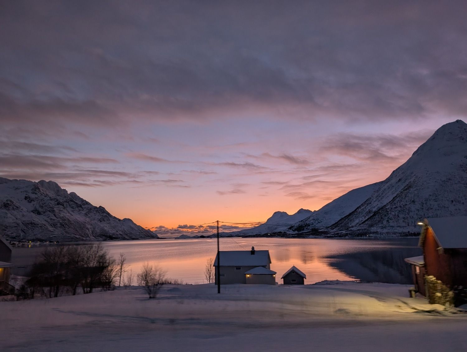 Un paysage hivernal paisible en Norvège avec des maisons enneigées au bord d'un fjord sous un ciel de coucher de soleil violet et orange.
