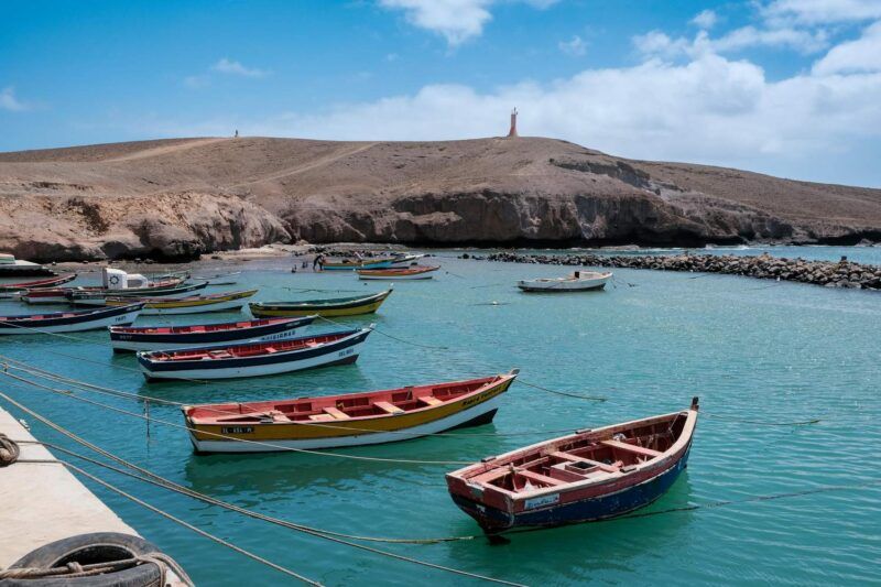 Barques de pêche colorées amarrées dans une baie d'eau turquoise face à une colline rocheuse surmontée d'une statue