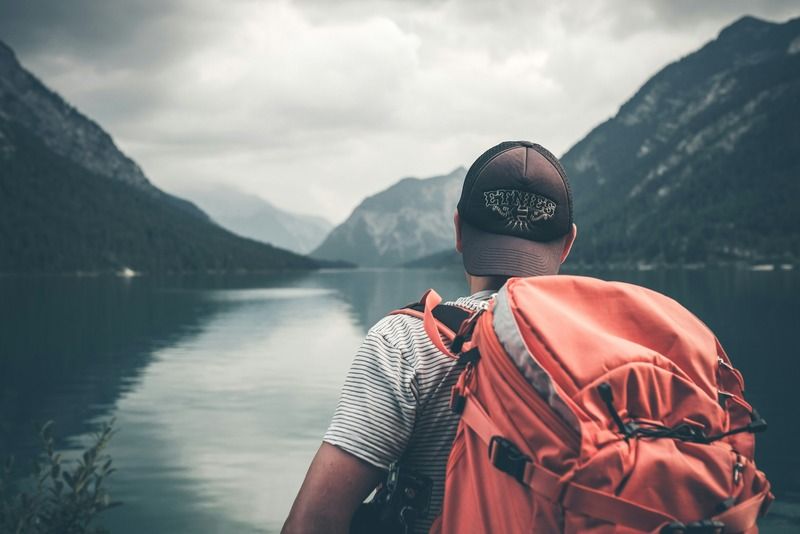 Randonneur avec un sac à dos orange contemplant un lac alpin entouré de hautes montagnes sous un ciel nuageux
