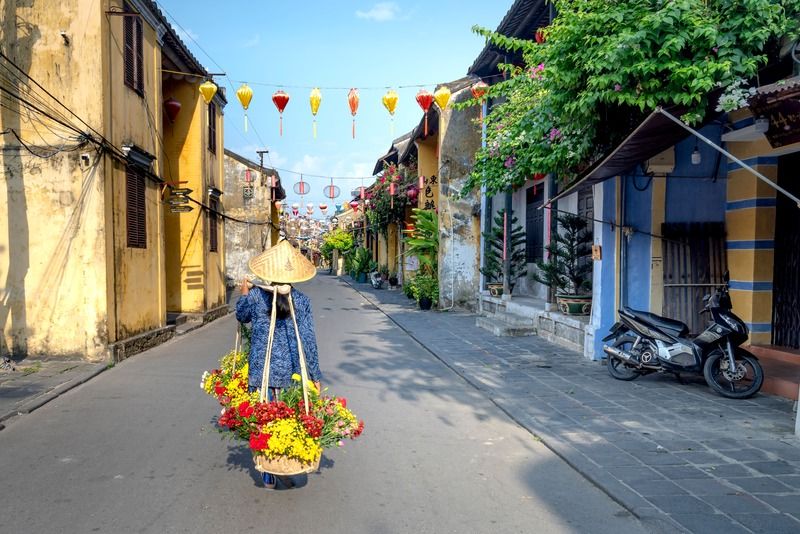 Rue colorée de la vieille ville de Hoi An avec des lanternes traditionnelles suspendues, des maisons jaunes et une vendeuse portant un chapeau conique, Vietnam