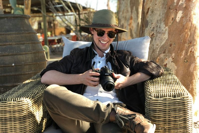 Homme souriant avec un chapeau et des lunettes de soleil tenant un appareil photo, assis sur une terrasse en rotin à l'extérieur
