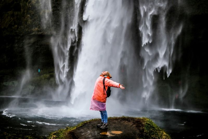 Femme en imperméable rouge les bras ouverts devant une grande cascade, sac à dos au dos