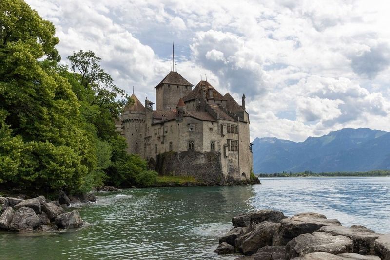 Le château de Chillon au bord du lac Léman, entouré de rochers et de verdure, avec les Alpes en arrière-plan