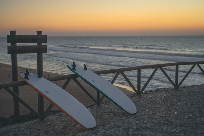 Deux planches de surf appuyées contre une barrière en bois face à l'océan, panneau « São Julião » au coucher du soleil