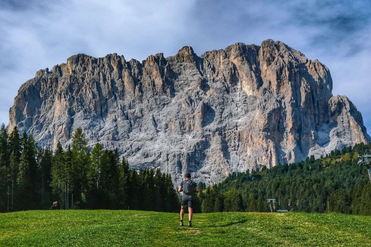 Randonneur dans une prairie face à une grande paroi rocheuse bordée de sapins