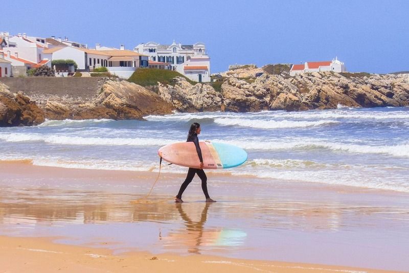 Surfeuse marchant sur la plage avec sa planche, village blanc portugais perché sur la falaise en arrière-plan