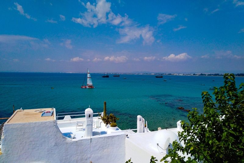 Terrasse blanche typique avec petit dôme, cactus et voilier voguant sur la mer turquoise à Hammamet