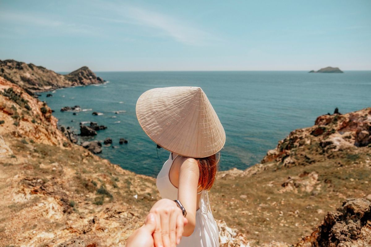 Femme au chapeau conique tenant une main face à une côte rocheuse et à la mer