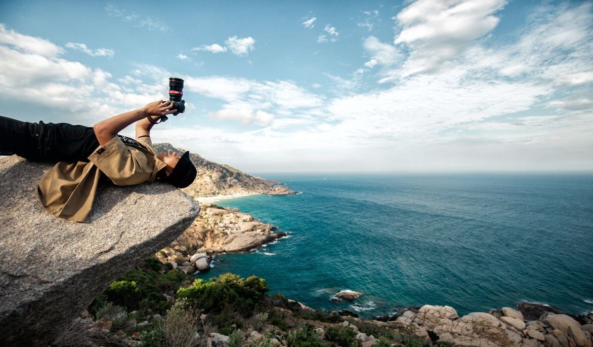 Photographe allongé sur un rocher levant son appareil face à la mer