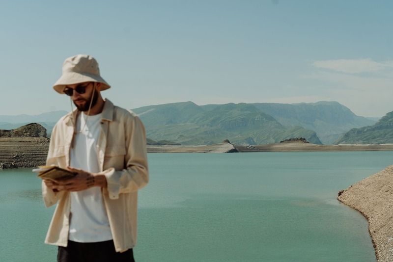 Un voyageur avec un chapeau de pêcheur et des lunettes de soleil lisant un carnet au bord d'un lac turquoise entouré de montagnes vertes