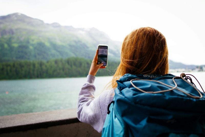 Voyageuse avec un sac à dos photographiant un lac alpin turquoise entouré de montagnes vertes